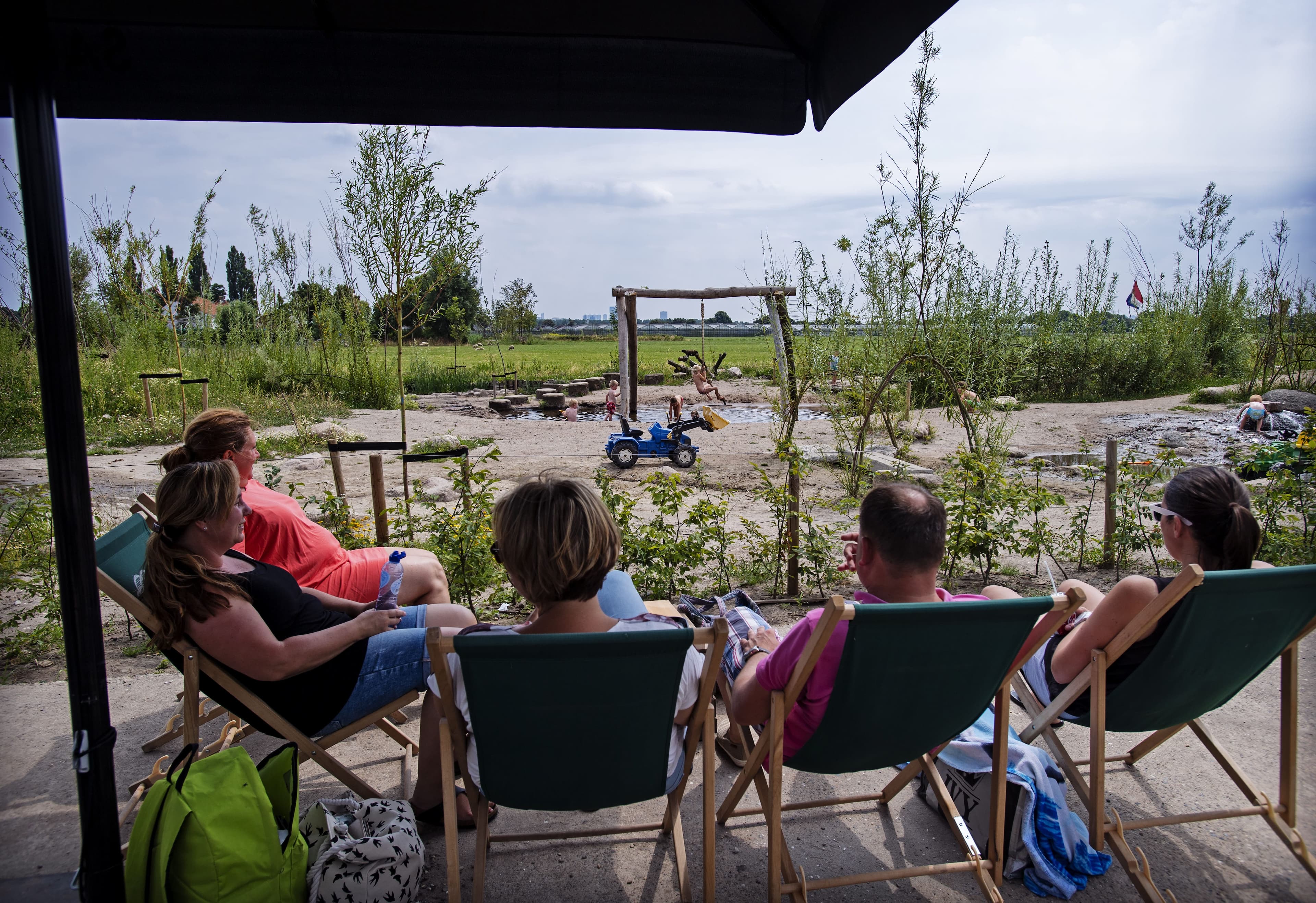 Vijf mensen zittend in strandstoelen kijkend naar spelende kinderen, in een natuurlijke speeltuin.